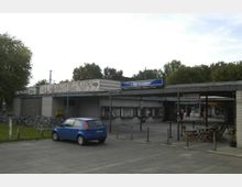 Außenansicht des U-Bahnhofs Hansaplatz mit flachem Eingangsgebäude aus hellem Backstein; links eine Fahrradabstellfläche, vorne ein blauer Kleinwagen. Neben dem Eingangsbereich stehen Tische und Stühle eines Cafés, im Hintergrund sind Bäume sichtbar.
