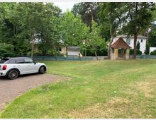 Eine grüne Rasenfläche mit einem weißen Auto, das auf einem gepflasterten Bereich geparkt ist. Im Hintergrund sind Bäume, ein kleines steinernes Pavillon mit rotem Ziegeldach und mehrere Einfamilienhäuser mit Zäunen zu sehen.