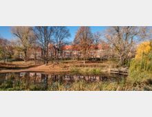 Ein Park mit einem kleinen Teich im Vordergrund, umgeben von Schilf und Bäumen mit herbstlichem Laub. Im Hintergrund ist ein langes Gebäude mit roten Ziegelwänden und Satteldach zu sehen, das von kahlen und spärlich belaubten Bäumen gesäumt wird.