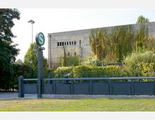 Eine S-Bahn-Station mit einem grünen „S“-Symbol auf einem Pfeiler im Vordergrund. Im Hintergrund ist ein großer, von Efeu bewachsener Betonbau zu sehen, umgeben von Bäumen und Sträuchern.