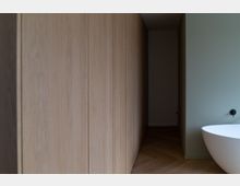 Minimalistisches Badezimmer mit Holzwänden auf der linken Seite, hellem Fischgrätboden und einer freistehenden weißen Badewanne vor einer hellgrünen Wand.