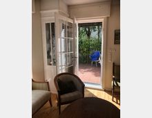 Wohnzimmer mit Holzboden, mehreren gepolsterten Sesseln und einem runden Holztisch, das durch eine geöffnete Glastür auf eine Terrasse mit Terrakottafliesen führt. Im Außenbereich sind grüne Pflanzen sowie ein blauer Stuhl sichtbar.