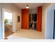 Flur mit heller Holzboden, beidseitig flankiert von Türen; an der rechten Wand befindet sich eine offene Garderobe mit orangefarbenen Vorhängen und Kleiderstange. Im Hintergrund ist ein angrenzender Raum mit farbigen Stühlen und Holzboden sichtbar.