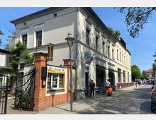 Ecke eines historischen Gebäudes mit weißer Fassade und verzierten Fenstern; rechts ein Gehweg mit Passanten und Kopfsteinpflasterstraße. Neben dem Eingang befindet sich ein Ladenfenster mit gelbem Schild „bee boy“, davor schwarze Zäune und ein gemauerter Torpfosten aus rotem Backstein.