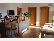 Heller Innenraum mit Holzboden, bestehend aus einem Essbereich mit Holztisch und einer kleinen Theke aus Holz mit Barhocker. Im Hintergrund befinden sich drei Türen mit Holzrahmen, eine an der Wand montierte TV-Bildschirm und mehrere Zimmerpflanzen.