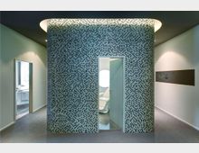 Runder Wandbereich mit Mosaikfliesen und eingelassenem Türdurchgang, der in einen Raum mit weißem Behandlungsstuhl und einer runden Wandbeleuchtung führt; rechts und links befinden sich offene Durchgänge zu weiteren Räumen.