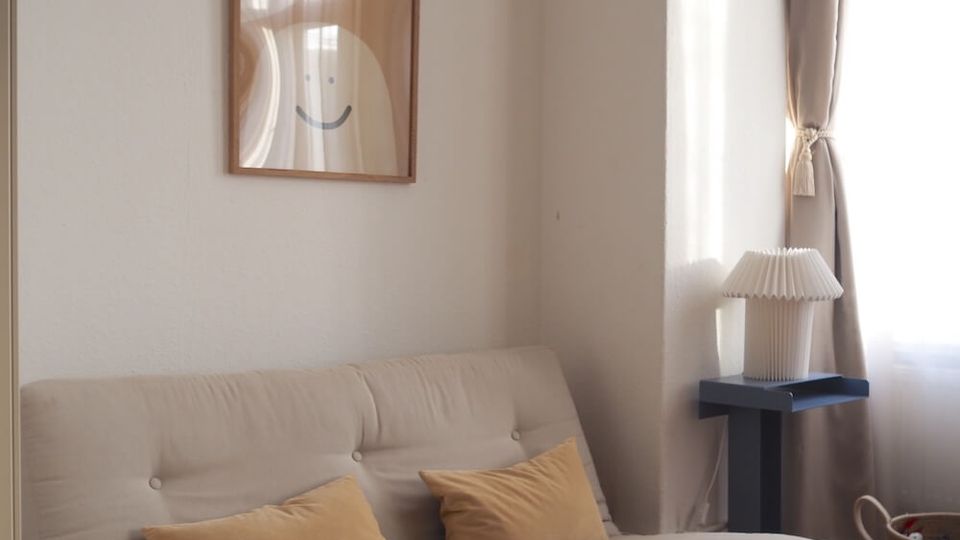 Bright living room corner with a beige tufted futon sofa on a wooden frame and two tan pillows, beneath a framed abstract print on a white wall. A tall window with sheer curtains and drapes lets in sunlight, with a small side table and pleated lamp beside the sofa on a wooden floor.