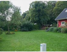 Ein grüner Garten mit einem roten Holzhaus am rechten Bildrand. Der Garten ist von Bäumen, Hecken und bunten Blumen umgeben, mit einem Rasen im Vordergrund und einem Stapel Brennholz im Hintergrund.