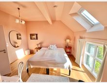 Helles Schlafzimmer mit Holzfußboden, pastellrosa Wänden und schräger Decke. Im Raum befinden sich ein Doppelbett, ein kleiner roter Nachttisch mit Tischlampe, ein runder Wandspiegel, sowie ein schräges Dachfenster und ein großes Fenster mit weißen Vorhängen.