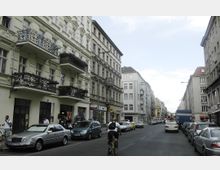 Straßenszene in einer Stadt mit Altbauwohnungen, verzierten Balkonen und Geschäften im Erdgeschoss. Autos parken am Straßenrand, während ein Fahrradfahrer die Straße entlangfährt.