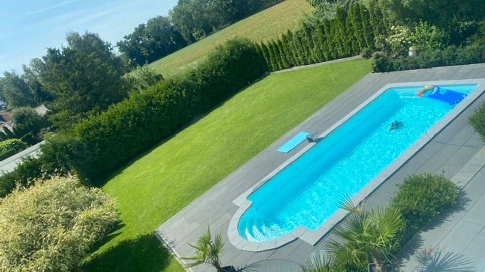 Rechteckiger Außenpool mit grauer Steinumrandung in einem gepflegten Garten mit großem Rasen. Umgeben von Hecken, Sträuchern und kleinen Palmen, mit Blick auf einen bewaldeten Hang und offene Wiesen im Hintergrund.