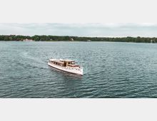 Ein weißes Boot mit Holzelementen fährt auf einem ruhigen See, umgeben von einer bewaldeten Uferlandschaft mit einzelnen Gebäuden im Hintergrund unter bewölktem Himmel.
