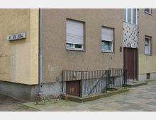 Mehrfamilienhaus mit bräunlicher Fassade, zwei Fenstern mit geschlossenen Rollläden und einem Hauseingang mit dunkelbrauner Tür. Im Vordergrund sind ein Gehweg, ein niedriger Metallzaun und Fahrradständer zu sehen.