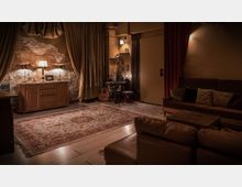 Gemütlich eingerichteter Raum mit warmem Licht, einer Kommode vor einer freigelegten Backsteinwand, flankiert von Vorhängen. Im Vordergrund ein orientalisch gemusterter Teppich, daneben ein Sofa mit Kissen; im Hintergrund stehen eine Akustikgitarre und eine kleine Trommel neben einem Tisch.