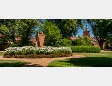 Gepflegter Stadtpark mit geschwungenen Sandwegen, großen Laubbäumen und einem runden Blumenbeet mit weißer Blüte um ein steinernes Denkmal. Im Hintergrund sind rote Dächer und ein Kirchturm mit grüner Kuppel zu sehen.