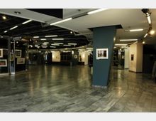 Großer, teilweise beleuchteter Innenbereich eines Gebäudes mit glatten, grauen Marmorböden; an den Wänden hängen eingerahmte Bilder. Im Hintergrund ist eine Garderobe ausgeschildert, und die Decke ist mit mehreren Leuchtstofflampen ausgestattet.