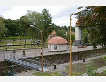 Blick auf eine Schleusenanlage mit einem kleinen, rundlichen Gebäude mit rotem Ziegeldach im Vordergrund. Umgeben von Lampenmasten, Bäumen und einer grünen Parklandschaft im Hintergrund.
