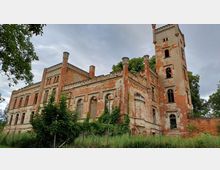 Verfallenes historisches Gebäude mit roter Backsteinfassade, teils zerstörten Fenstern und einer hohen, rechteckigen Turmstruktur. Das Gebäude ist von Gras und Bäumen umgeben, der Himmel ist bewölkt.