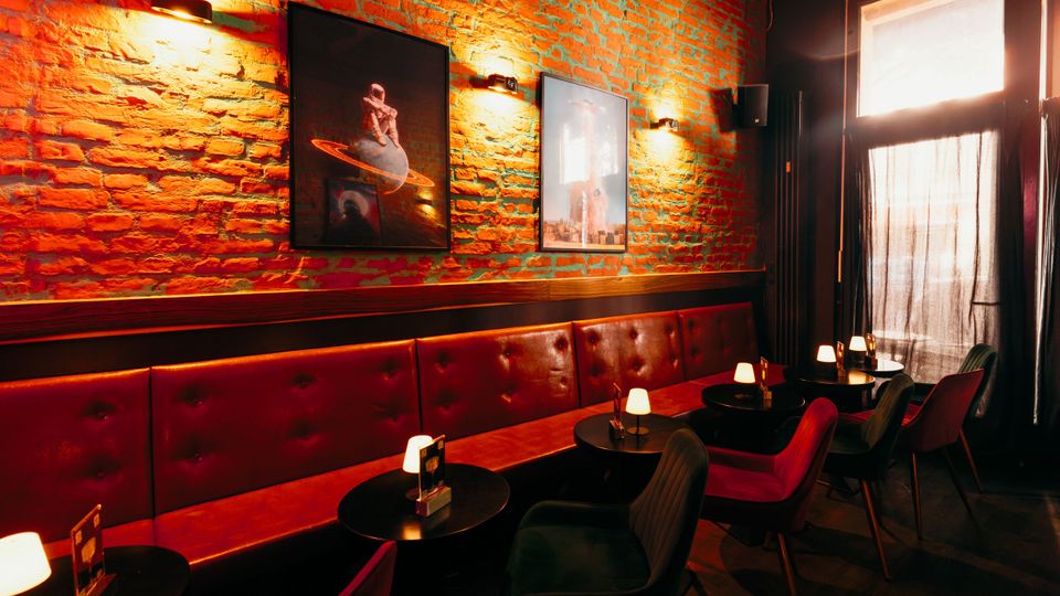 Innenraum einer Bar oder Lounge mit freiliegender Backsteinwand, gerahmten Bildern und warmen Wandleuchten über einer langen, gepolsterten roten Sitzbank. Davor stehen kleine runde Tische mit Tischlampen und dunkle Stühle; rechts fällt Licht durch ein großes Fenster mit Vorhang.
