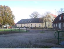 Hof mit mehreren Gebäuden in ländlicher Umgebung, darunter ein gelbes Hauptgebäude mit Schrägdach und ein steinernes Gebäude mit rotem Dach. Im Vordergrund ist ein gepflasterter Weg, ein Zaun und teils parkende Fahrzeuge, darunter ein Traktor, zu sehen.