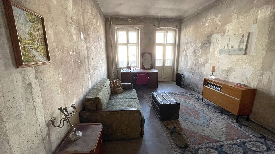 Langer, schmaler Wohnraum mit stark abgenutzten, fleckigen Wänden und hoher Decke, eingerichtet mit Sofa, Teppich, Truhe als Couchtisch, Sideboard und kleinem Schreibtisch am Fenster. Am Ende des Raums lassen zwei hohe Fenster und eine verglaste Tür viel Tageslicht herein.