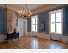 Eleganter Raum mit blauen Wänden, verziert mit weißen und goldenen Ornamenten, einem Kronleuchter, großen Fenstern mit weißem Rahmen und einem aufwendig gestalteten Kamin. Der Boden ist aus hellem Parkett, und ein vergoldeter Wandspiegel hängt an der rechten Wand.