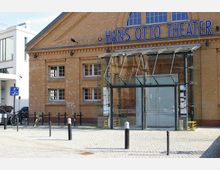Fassade des Hans Otto Theaters, ein Gebäude aus gelbem Klinkerstein mit großen Fenstern und einem modernen gläsernen Eingangsbereich. Im Vordergrund ist eine gepflasterte Fläche mit Fahrrädern und Pollern zu sehen.