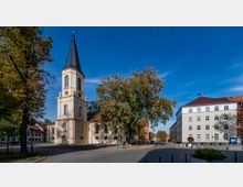 Historische Kirche mit hellem Turm und spitzem Dach auf einem gepflasterten Platz, umgeben von Bäumen und angrenzenden Gebäuden mit roten und hellen Fassaden unter klarem blauen Himmel.