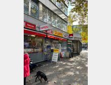 Straßenansicht eines kleinen Ladengeschäfts mit Lotto-Annahmestelle, Zeitschriften und Zeitungen im Schaufenster. Über dem Geschäft befinden sich große Fenster eines Büro- oder Wohngebäudes, umgeben von Bäumen mit herbstlichem Laub.