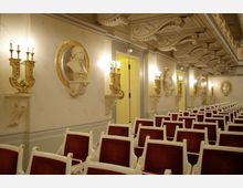 Eleganter Konzertsaal mit roten Polsterstühlen und verzierten weißen Rahmen. An den Wänden befinden sich goldene Wandlampen in Form von Kandelabern sowie Marmorbüsten in goldenen Rahmen, die von kunstvoll gestalteten Stuckelementen umgeben sind.