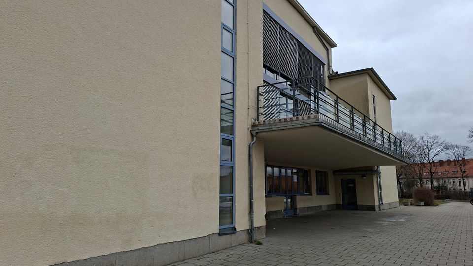 Außenansicht eines modernen, beigefarbenen Gebäudes mit großen Fenstern, einem überdachten Eingangsbereich und einem auskragenden Balkon mit Metallgeländer. Davor liegt ein gepflasterter Vorplatz; im Hintergrund sind Bäume und weitere Wohnhäuser zu sehen.