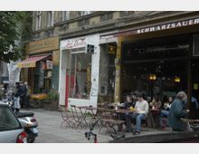 Straßenszene mit mehreren kleinen Geschäften und einem Café in Berlin, darunter ein Laden mit der Aufschrift "Koof im Kiez!" und ein Café mit roten Tischen und Stühlen im Außenbereich, wo Menschen sitzen und speisen. Die Fassade der Gebäude ist alt und teils mit Graffiti bedeckt.