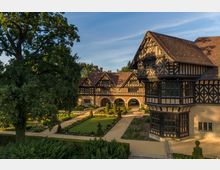 Historisches Herrenhaus im Fachwerkstil mit roten Ziegeldächern und verzierten Erkern, umgeben von einem gepflegten Garten mit Rasenflächen, Kieswegen und Hecken. Im Hintergrund sind Bögen und ein großer Baum zu sehen, der Schatten über einen Teil des Gartens wirft.