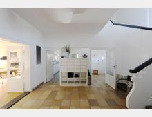 Heller Eingangsbereich mit beigem Fliesenboden, weißen Wänden und einer modernen weißen Treppenkonstruktion rechts. In der Mitte des Raumes steht ein weißes Regal, links führt eine Glastür zu einem Büro mit Tisch und Stühlen.