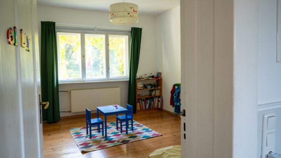 Blick durch eine offene Tür in ein helles Kinderzimmer mit Holzfußboden, großer Fensterfront und grünen Vorhängen. In der Mitte stehen ein kleiner blauer Kindertisch mit zwei Stühlen auf einem bunten Teppich; rechts befindet sich ein niedriges Bücherregal.