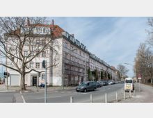 Wohnstraße mit mehrstöckigem Wohngebäude in heller Putzfassade und roten Ziegeldächern. Im Vordergrund verläuft eine Straße mit geparkten Autos, daneben Bürgersteige mit Bäumen, von denen einige unbelaubt sind.