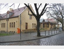 Ein einstöckiges Gebäude aus gelbem Stein mit Satteldach und mehreren Fenstern, umgeben von einem gepflasterten Gehweg und kahlen Bäumen. Die Straße im Vordergrund besteht aus Kopfsteinpflaster, und rechts ist ein überdachter Eingangsbereich zu sehen.