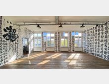 Heller Raum mit Holzboden und großen Fenstern, die Tageslicht hereinlassen. Die Wände sind vollständig mit großflächiger, handgeschriebener schwarzer Kalligrafie auf weißem Hintergrund bedeckt.