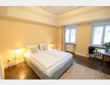 Helles Schlafzimmer mit zwei zusammengestellten Einzelbetten, Nachttischen mit Lampen und hohen Decken mit Stuck. Zwei große Fenster mit Vorhängen und Heizkörpern darunter sowie ein kleiner Tisch mit Stuhl stehen auf dunklem Fliesenboden mit hellem Teppich.