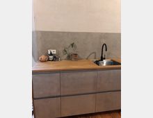 Moderne Küche mit einer Arbeitsplatte aus Holz, einer Spüle aus Edelstahl mit schwarzem Wasserhahn und grauem Unterschrank. Im Hintergrund eine Wand mit Betonoptik und darauf eine Pflanze in einem Blumentopf sowie kleine Küchenutensilien.