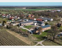Luftaufnahme eines ländlichen Gutshof- und Dorfbereichs mit mehreren langgestreckten Wirtschaftsgebäuden und roten sowie grauen Dächern, umgeben von Wiesen und Feldern. Im Hintergrund sind ein kleiner Ortskern mit Kirchturm, Baumgruppen und weitläufige Ackerflächen bis zum Horizont zu sehen.