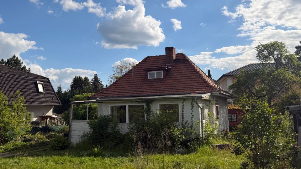 Einfamilienhaus mit rotem Ziegeldach und Schornstein in einem verwilderten Garten mit hohem Gras und dichtem Bewuchs. Im Hintergrund stehen weitere Wohnhäuser und Bäume unter einem teils bewölkten Himmel.