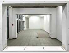 Ein leerer, rechteckiger Raum mit weißen Wänden und grauen Fliesenboden. Auf der linken Seite befinden sich große Fensterfronten mit Glastüren, ein Heizkörper hängt an der Rückwand, und die Decke ist weiß mit eingelassenen Leuchten.