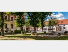 Ein kleiner Park mit Bäumen, Bänken und Blumenkübeln, in dessen Zentrum ein steinernes Denkmal mit einer Inschrift steht. Im Hintergrund sind ein historisches Wohngebäude mit verzierten Fassaden und ein weißes Gebäude mit roten Ziegeldächern zu sehen.