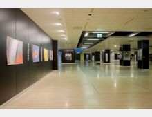 Eine moderne Ausstellungshalle mit hellen Holzböden, schwarzen Trennwänden und einer abgehängten Decke mit integrierter Beleuchtung. An den Wänden hängen verschiedene farbige Kunstwerke, und der Raum wirkt offen und geräumig.