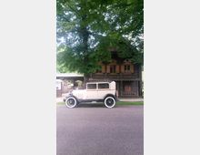 Ein cremefarbenes Oldtimer-Auto mit schwarzen Details steht vor einem rustikalen Holzhaus mit geschnitzten Verzierungen und geschlossenen Fensterläden, umgeben von grünen Laubbäumen.