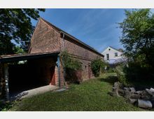 Backsteingebäude mit Satteldach und Holzläden, neben einem offenen Schuppen, umgeben von Bäumen und Sträuchern. Im Vordergrund befindet sich eine Wiese mit gestapelten Holzscheiten, im Hintergrund ein weiß verputztes Haus.