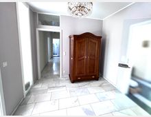 Heller Eingangs- oder Flurbereich mit hellgrauen Wänden und großformatigem, marmoriertem Fliesenboden. In der Ecke steht ein dunkler Holzschrank; darüber hängt ein Kristalllüster, daneben führt eine Tür mit Milchglas in den Raum und ein offener Durchgang in einen langen Flur.