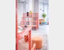 Heller, moderner Loft-Innenraum mit hohen Decken, weißen Wänden und Vorhängen sowie sichtbaren Installationen an der Decke. In der Mitte steht ein rotes Metallregal mit Holzablagen; daneben ein Sitzbereich mit orangefarbenem Sofa, blauem Couchtisch, Teppich, Hocker und einer großen Zimmerpflanze unter Pendelleuchten.