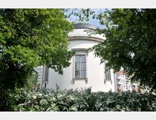 Rundbau einer weißen Kirche mit großen Rundbogenfenstern, umgeben von blühenden Sträuchern und hohen Bäumen; im Hintergrund sind Wohngebäude sichtbar.